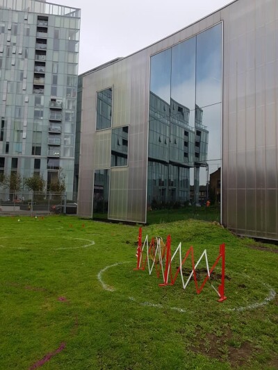 A piece of red and white fencing marking a man-hole in the middle of a white circle on the grass in front of Trinity Laban's Creekside building