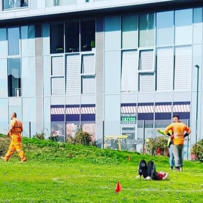 Dance student lying down on the grass with three people  in hi-vis work outfits behind them