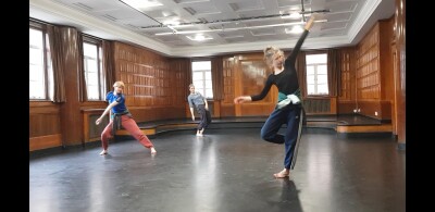 Three dancers in a wood panelled studio with a black floor.  They make angular shapes with this bodies