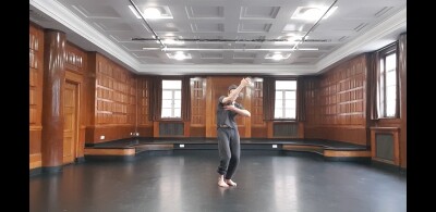 A man dressed in black sweats and t shirt holds his body in an s-shape with his right arm raised and obscuring his face while his left hand touches his chest.  He's in the middle of a wooden panelled studio with a black floor and white panelled ceiling.