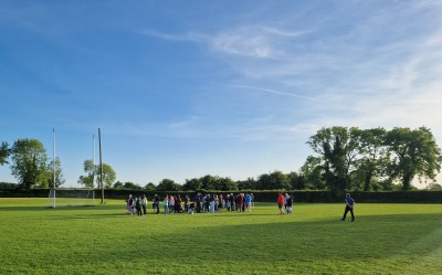 Approximately fifty people of all ages, children to older adults, gather on a green GAA pitch with a blue sky overhead and evening sunlight shining through the large trees on the pitch's perimeter