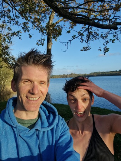 Fearghus and Isabella selfie after a swim in the lake seen behind them.  They're under a tree.  He's wearing a blue hoodie and smiling,  She's wearing a bikini top and has a hand raised to her short hair. The sunlight is shining in their faces