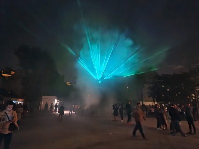 Image of a dark courtyard with figures walking through smoke.  The sky above is lit with blue lasers
