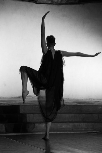 A dancer in a black silk garment balances on her right leg with left knee raised high, left arm above their head and right extended straight to the side.  They face away from camera to a while wall