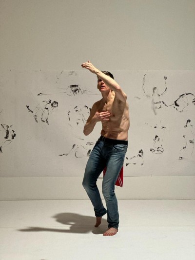 Fearghus topless in jeans against a white wall with charcoal drawings.  one arm is raised covering his face. the other hand touching his chest.