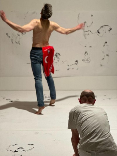 Gabriel in white crouching in foreground drawing Fearghus in background with arms wide and on tip toes.  Fearghus is topless, wearing blue jeans and with a red Cork hurling jersey hanging from his waist.  White background and floor with charcoal drawings