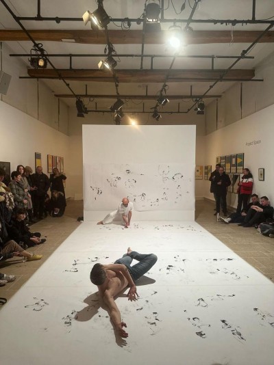 And exhibition space with people om the sides watching Fearghus lying on the ground in the foreground and Gabriel crouching and drawing in the background.  White back wall and ground  covered in charcoal drawings