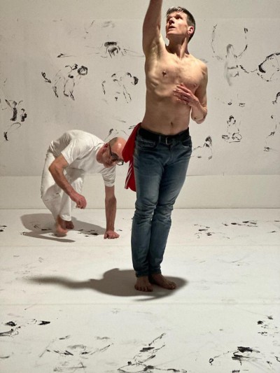 White background and floor covered in charcoal drawings.  Gabriel crouching in bacjkgound in white t-shirt and trousers watching Fearghus topless and wearing blue jeans.  Fearghus is standing tall with one arm reaching above his head.