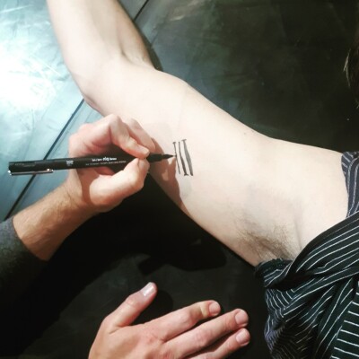 Image of inside of right forearm against a black floor with another pair of hands drawing with a pen an ink tattoo of three lines 