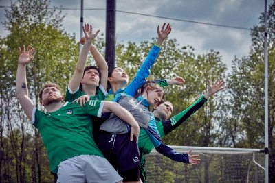 Five dancers in GAA kit of blue, green and black with arms extended as if reaching to catch a ball.  In the background trees, a cloudy sky and partially seen cross bar.