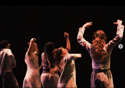 Four dancers in grey tone costumes against a back background.  Arms raised with bodies upright and twisting in motion