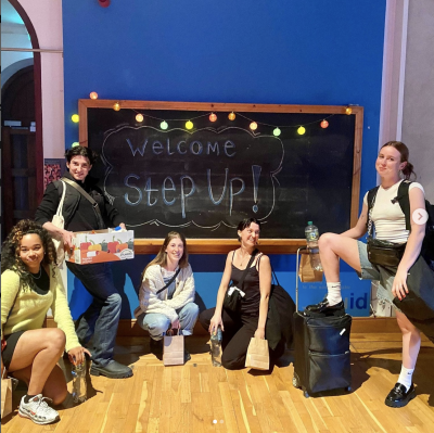 Five dancers pose with cheeky smiles in front of a blackboard that reads Welcome Step Up 