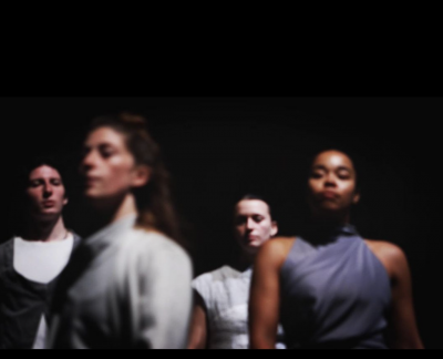 Soft focus photo of four dancers seen from chest up in grey tone costumes against a black background