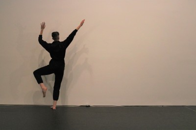 Dancer in black facing away from camera with hands raised and balancing on one leg with the other bent.  Against a white wall