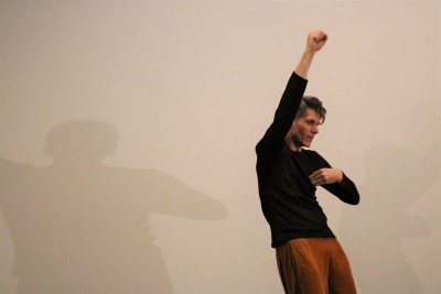 A man with right and raised and left hand touching his chest. He's wearing a dark sparkly top and a velour gold trousers. His shadow is visible on the white wall behind.