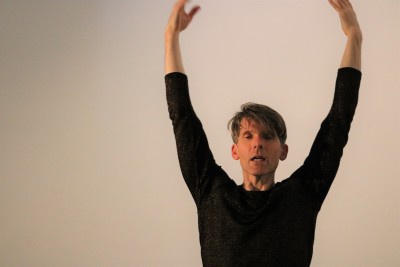 Male dancer with eyes closed and both arms raised over his head against a white background.