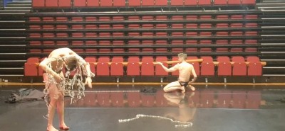 Two dancers in black briefs.  One on their knees upstage gesturing with his left hand to the left.  The other in the left hand corner downstage curved over with a garment of pearls falling over her.  A string of pearls on the ground between them